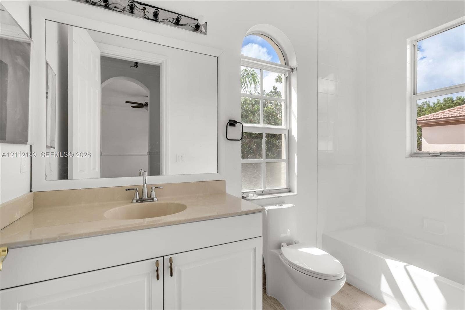 Bathroom featuring a white vanity cabinet, neutral countertop with integrated sink, arched window, and a built-in tub with tile surround
