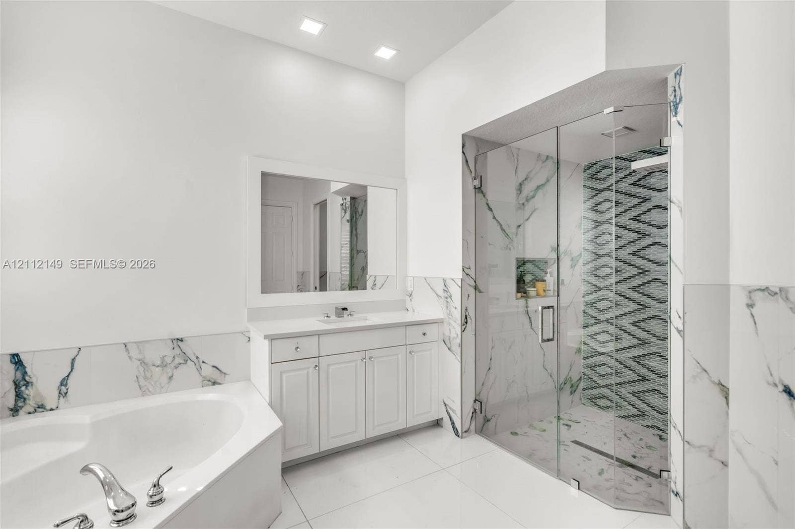 White vanity with integrated basin, framed wall mirror, soaking tub with deck-mounted faucet, walk-in shower with glass enclosure, and patterned tile accents