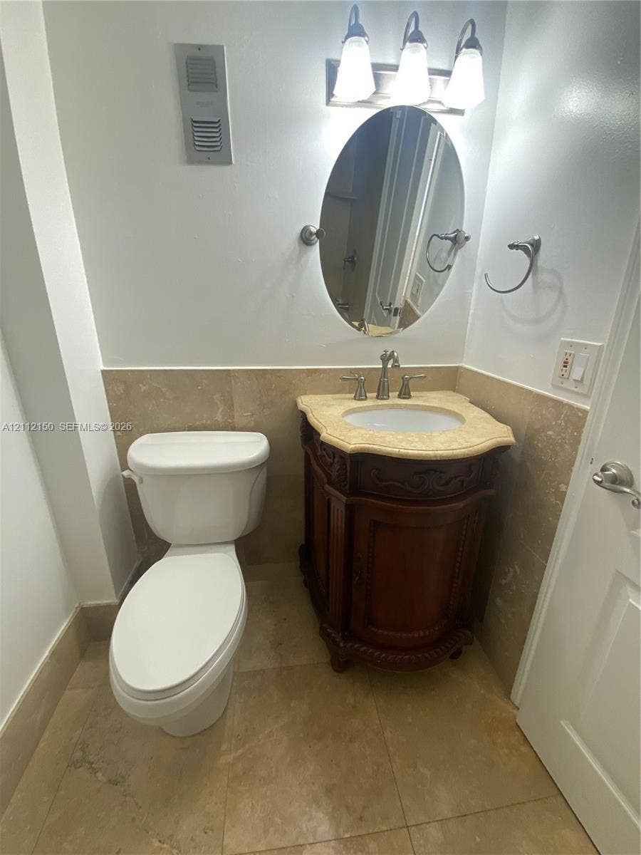 Custom wood-finish vanity with a stone countertop, oval undermount sink, three-light fixture, and neutral stone tile flooring and wall wainscoting