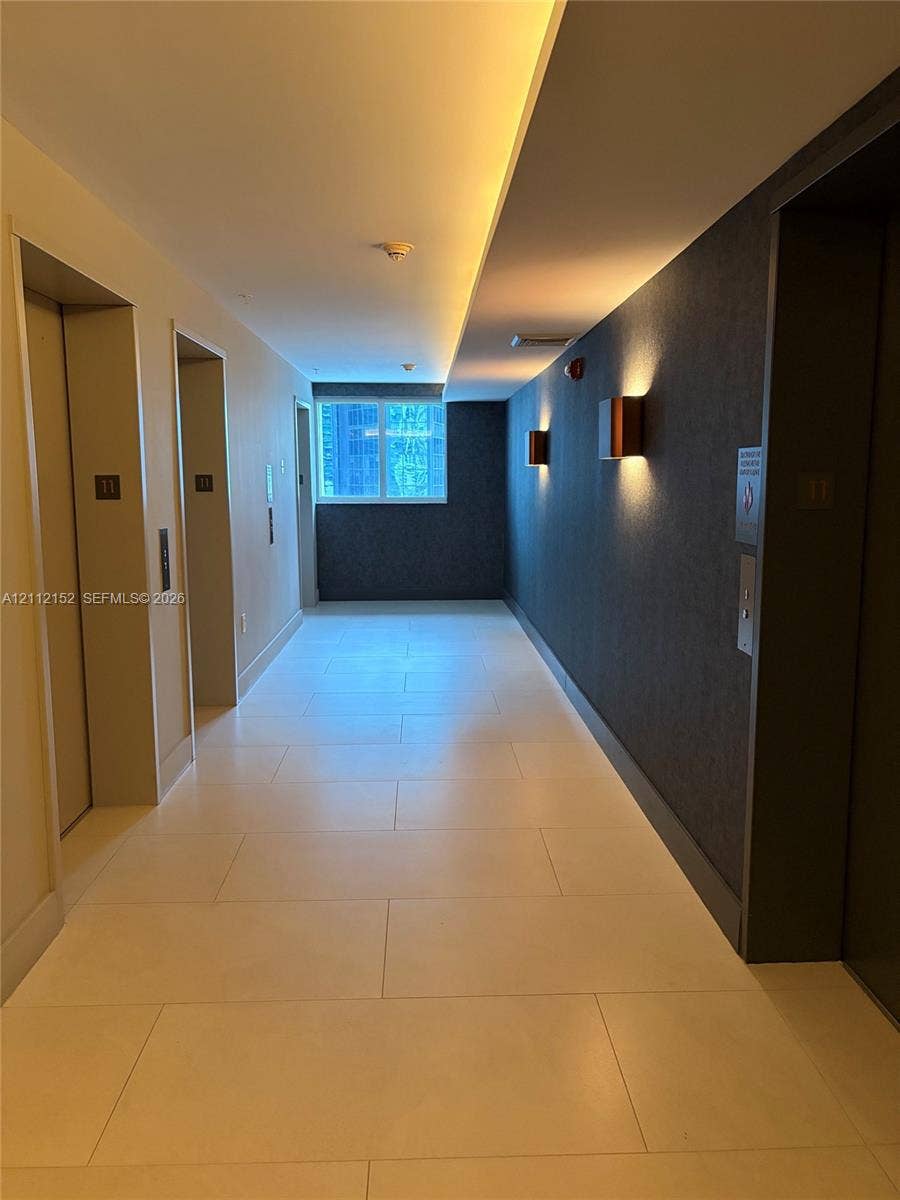 Hallway featuring large-format tile flooring, textured dark accent wall, recessed cove lighting, and metal-finished elevator banks