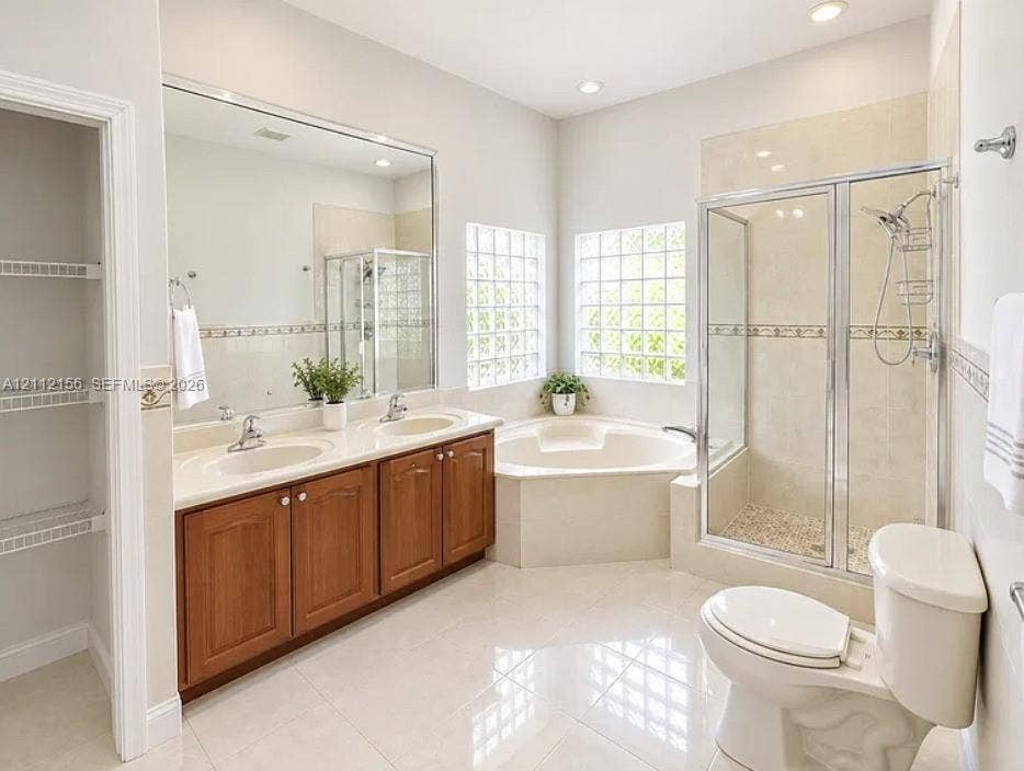Dual-sink vanity with wood-finish cabinetry, corner soaking tub, glass-enclosed shower, glass block windows, and built-in linen shelving