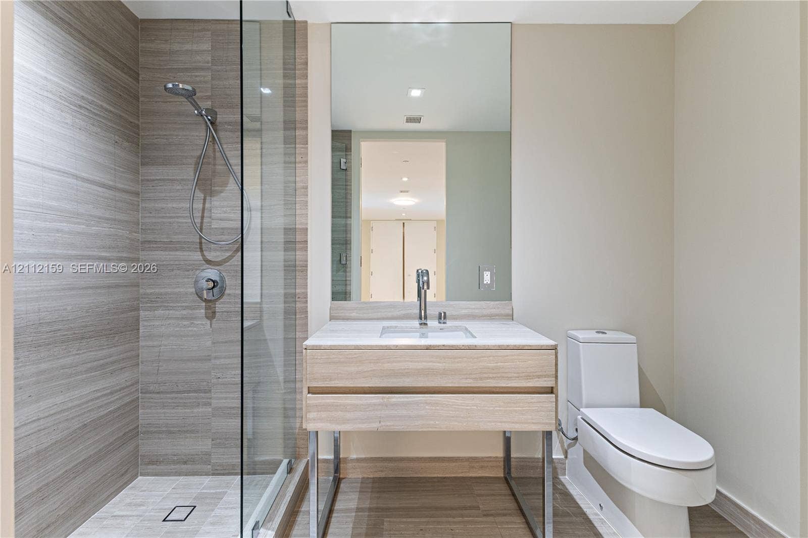 18975 Collins Avenue, #802, Sunny Isles Beach, Sunny Isles Beach, FL 33160 Modern bathroom featuring a wood-finish floating vanity with a light stone countertop, a glass-enclosed shower with horizontal-patterned wall tile, and a wall-to-wall vanity mirror