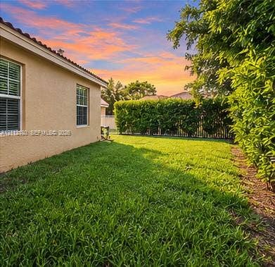 Backyard featuring a stucco exterior wall, dual-pane windows, green grass, mature perimeter hedges, and an enclosure fence