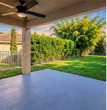 Covered patio featuring a ceiling fan, concrete flooring, manicured lawn, metal fencing, and perimeter hedging
