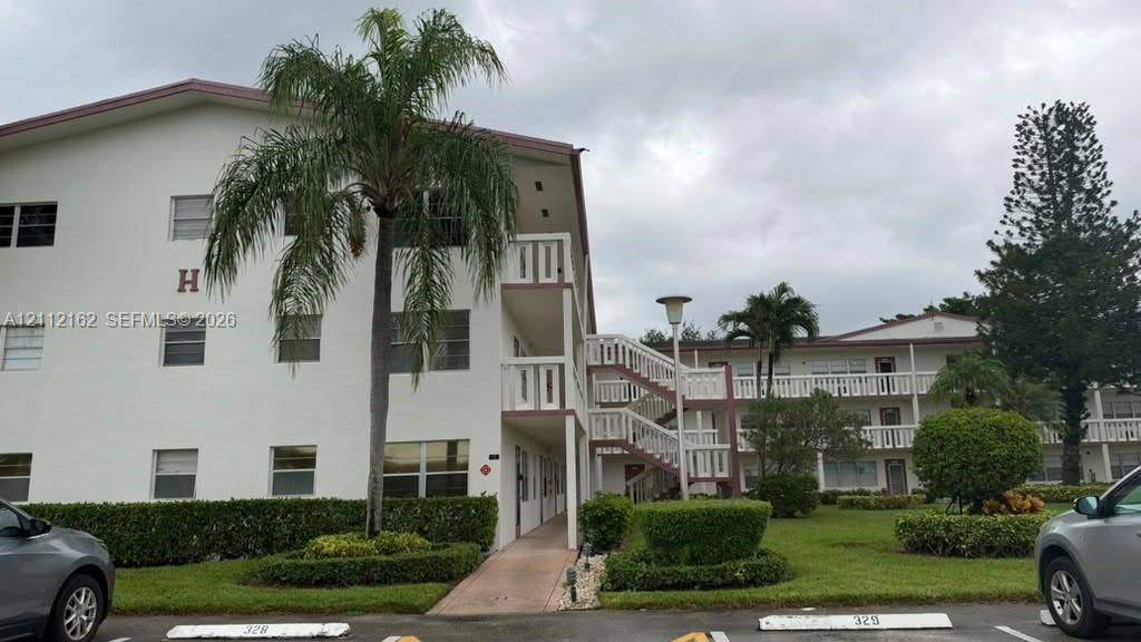309 Fanshaw H, #309, Boca Raton, FL 33434 Multi-story building exterior featuring white stucco siding, open-air walkways with balustrade railings, manicured shrubbery, and mature palm trees
