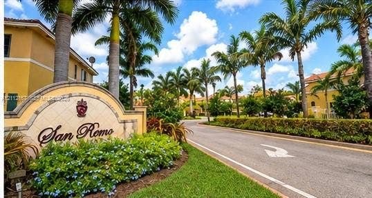 Community entrance featuring a stone-finish monument sign, mature palm trees, manicured landscaping, and asphalt paving