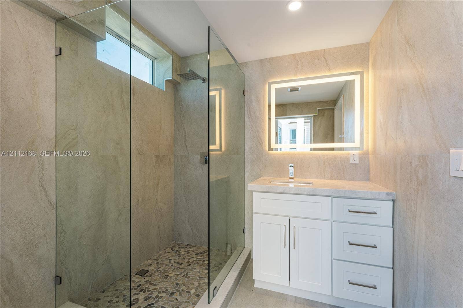 1041 E Buttonwood Drive, Sugarloaf Key, FL 33042 Walk-in shower with a glass enclosure, pebble tile floor, and rainfall showerhead, complemented by a white vanity with a stone countertop, integrated sink, and an illuminated mirror