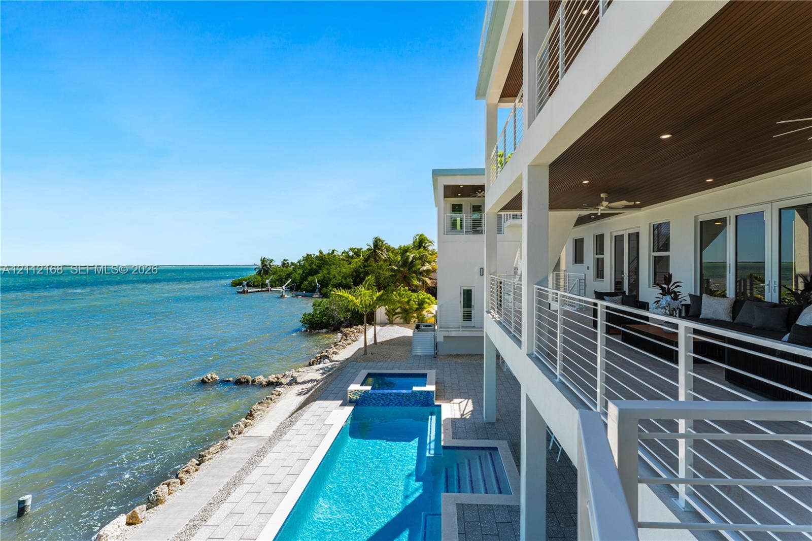 1041 E Buttonwood Drive, Sugarloaf Key, FL 33042 Multi-level waterfront residence featuring a lap pool with an integrated spa, expansive covered balcony with slatted wood-finish ceilings, metal railings, and stone-paver hardscaping