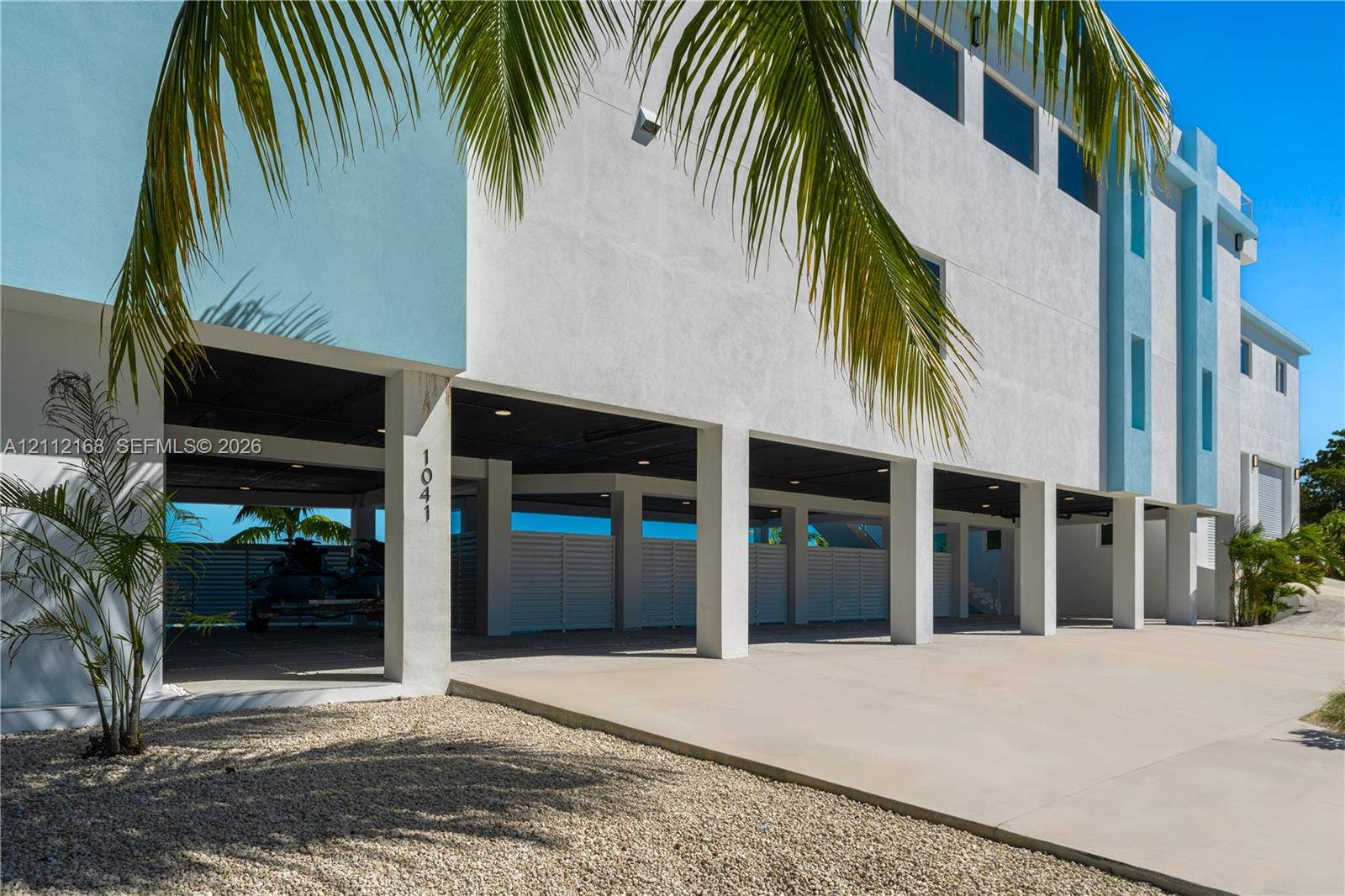 1041 E Buttonwood Drive, Sugarloaf Key, FL 33042 Modern multi-level exterior featuring white stucco finish, covered parking area, concrete driveway, horizontal slat privacy fencing, and recessed lighting