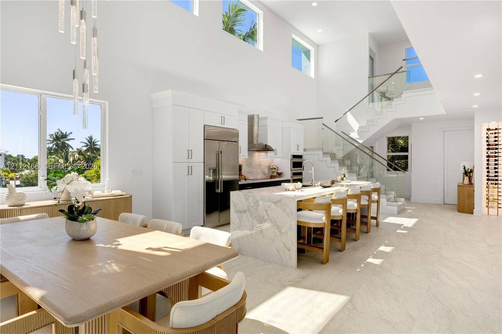 1041 E Buttonwood Drive, Sugarloaf Key, FL 33042 Open-concept kitchen featuring an expansive stone-topped waterfall island, custom white cabinetry, stainless steel professional-grade appliances, floating staircase with glass railings, and double-height ceilings with transom windows