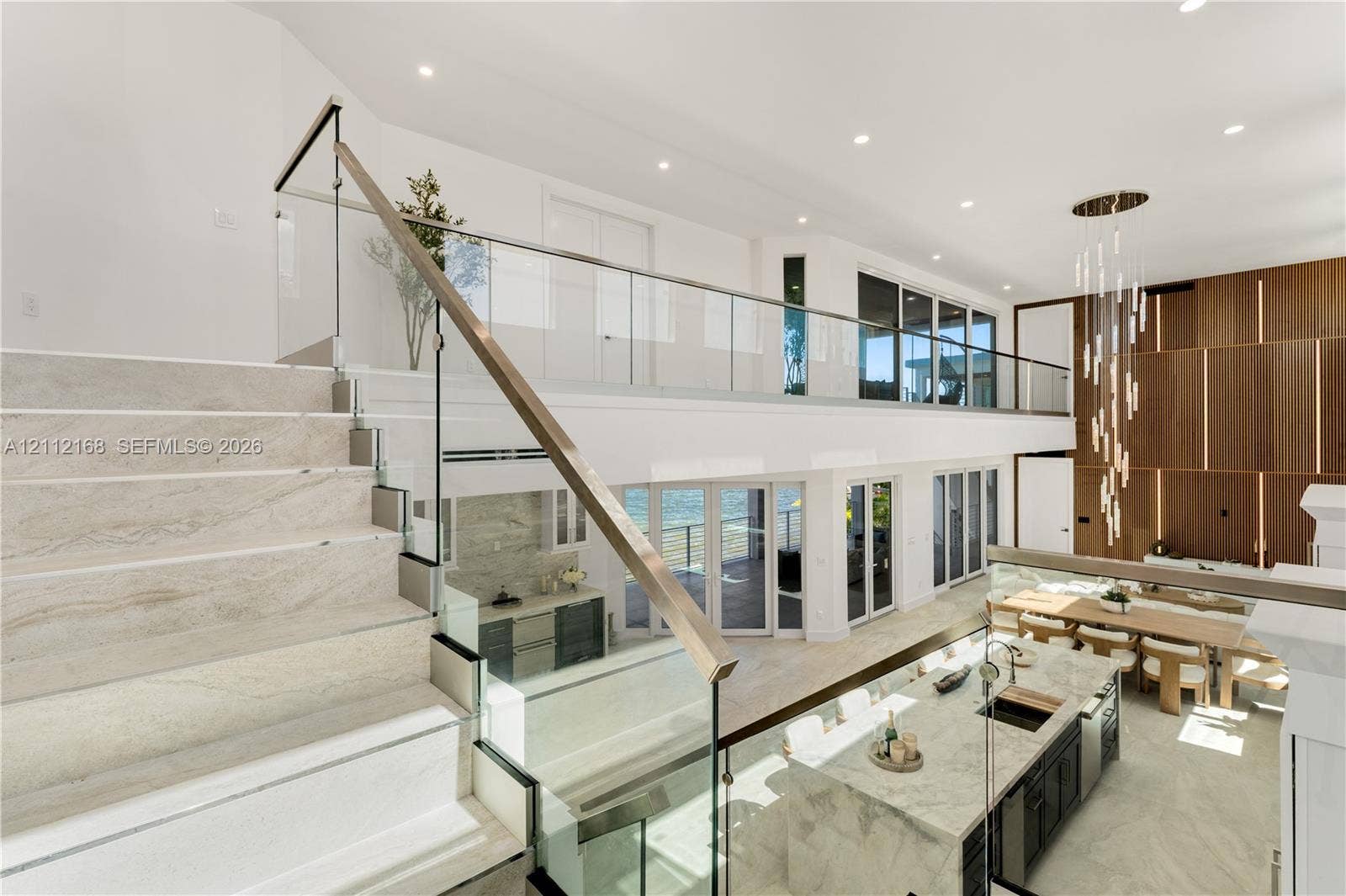 1041 E Buttonwood Drive, Sugarloaf Key, FL 33042 Open-concept interior featuring stone-finish flooring, a glass and metal staircase railing, a multi-level wood-panel wall feature, a stone-topped kitchen island, and multiple glass door systems