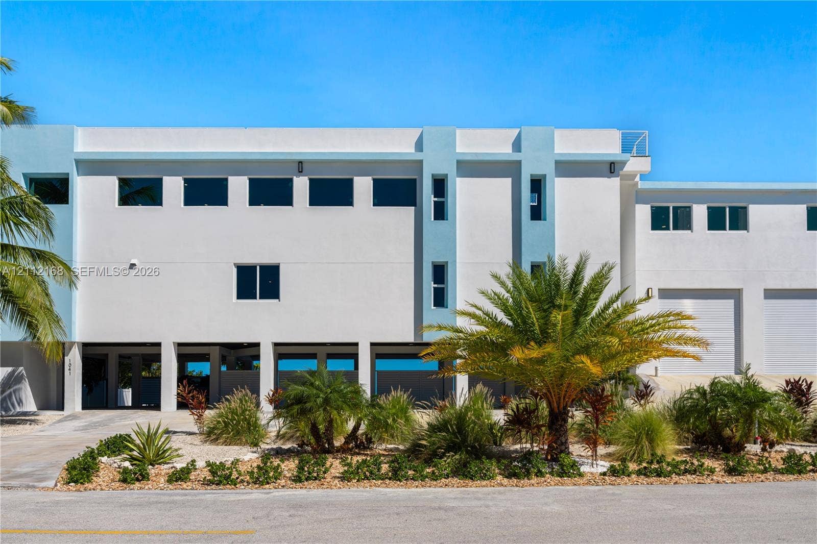 1041 E Buttonwood Drive, Sugarloaf Key, FL 33042 Multi-level exterior featuring smooth stucco finish, tiered flat roofline, oversized rectangular windows, ground-level covered parking, and attached multi-bay garage