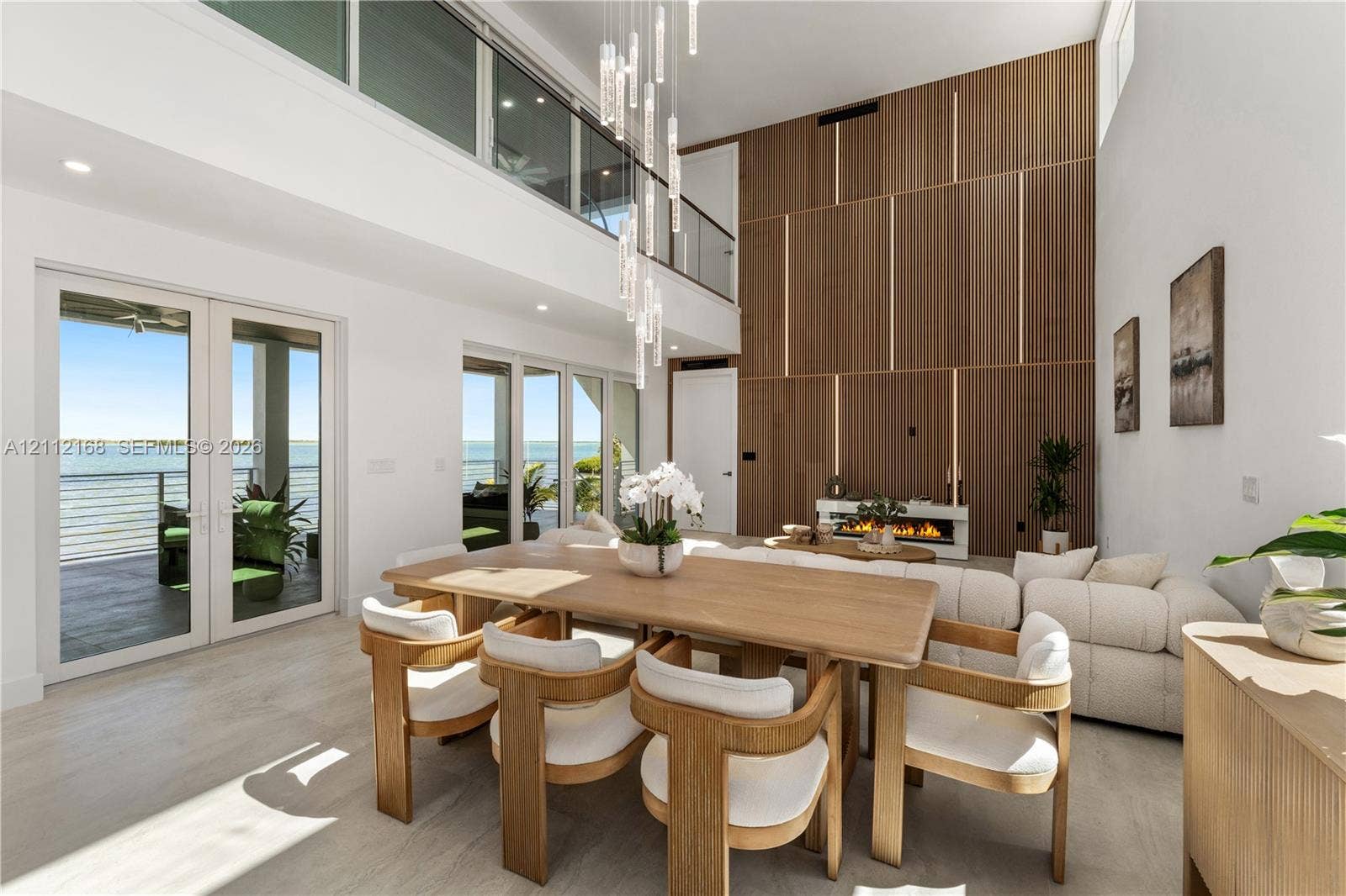 1041 E Buttonwood Drive, Sugarloaf Key, FL 33042 Double-height living area featuring floor-to-ceiling wood paneled accent wall with integrated linear lighting, glass-railed mezzanine, contemporary multi-pendant light fixture, and multiple sets of glass panel doors
