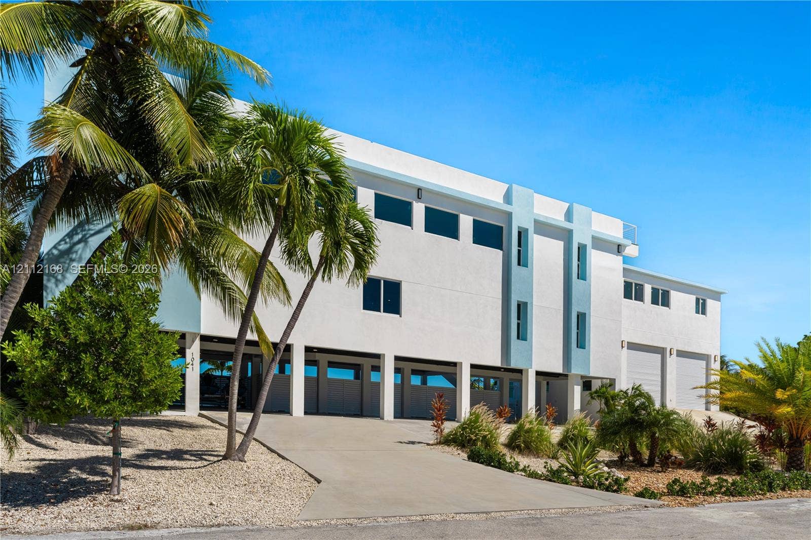 1041 E Buttonwood Drive, Sugarloaf Key, FL 33042 Contemporary multi-level exterior featuring white stucco finish, elevated structural design, integrated garage bays, and mature palm trees