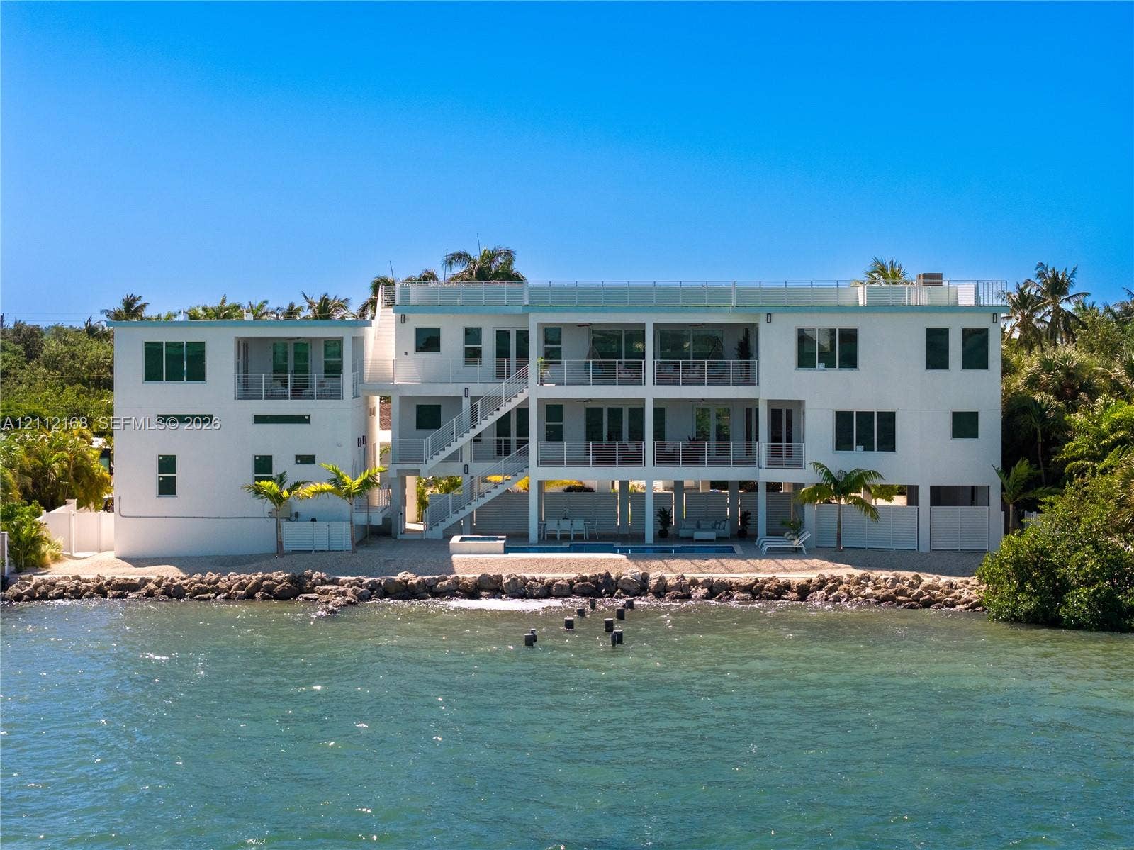 1041 E Buttonwood Drive, Sugarloaf Key, FL 33042 Multi-level waterfront structure featuring a white stucco exterior, tiered balconies with metal railings, an integrated ground-level swimming pool, and stone bulkhead shoreline reinforcement