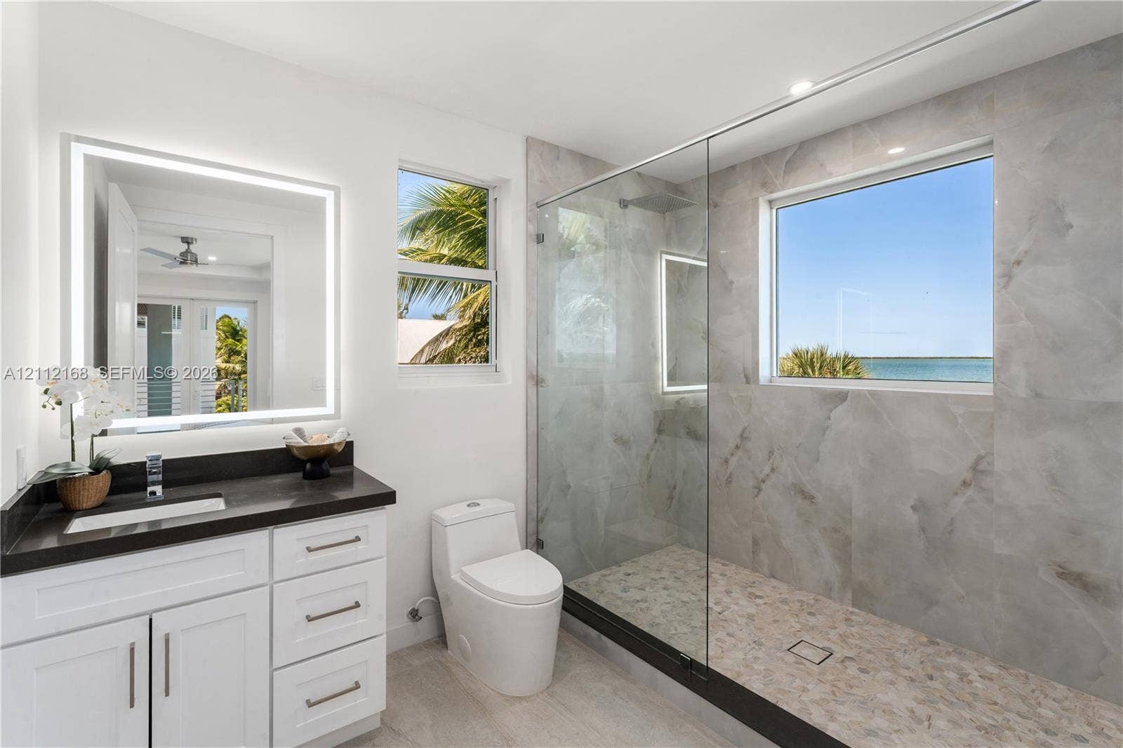1041 E Buttonwood Drive, Sugarloaf Key, FL 33042 Bathroom featuring a white vanity with a dark countertop, an illuminated mirror, a glass-enclosed shower with large-format tile surround, and an integrated window