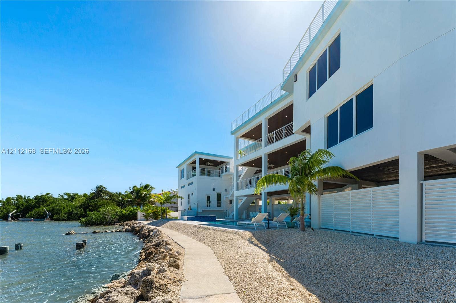 1041 E Buttonwood Drive, Sugarloaf Key, FL 33042 Multi-level waterfront exterior featuring white stucco siding, glass-panel railings, covered balconies, an inground swimming pool, and stone-lined shoreline