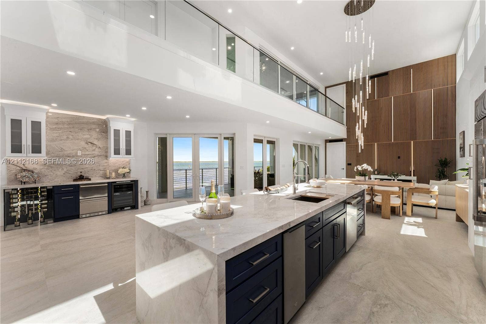 1041 E Buttonwood Drive, Sugarloaf Key, FL 33042 Double-height ceilings with custom wood-panel wall feature, oversized kitchen island with stone waterfall countertop, integrated floor-to-ceiling glass panel balcony railing, contemporary tiered pendant lighting, and multiple sets of glass exterior doors