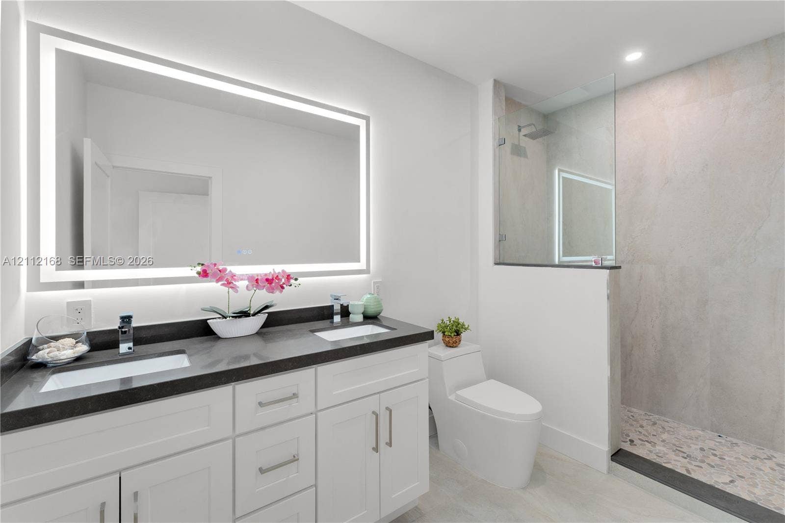 1041 E Buttonwood Drive, Sugarloaf Key, FL 33042 Double vanity with dark countertops, integrated rectangular sinks, wall-to-wall LED backlit mirror, walk-in glass shower with stone-style tile surround, and tile flooring