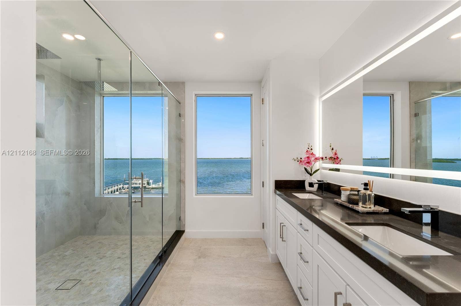 1041 E Buttonwood Drive, Sugarloaf Key, FL 33042 Dual-vanity bathroom featuring white cabinetry, dark stone countertops, a large frameless glass-enclosed shower with stone tile flooring, and a full-length illuminated vanity mirror