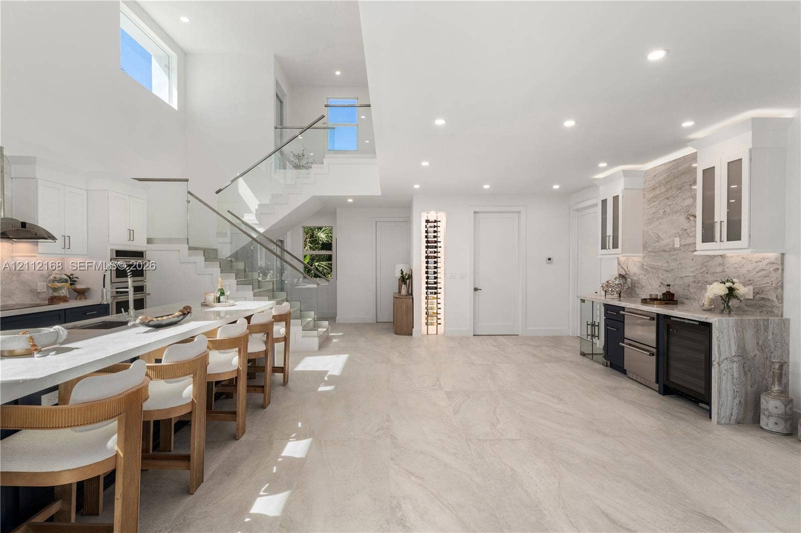 1041 E Buttonwood Drive, Sugarloaf Key, FL 33042 Open-concept kitchen featuring a large central island with waterfall-edge countertop, natural stone-style backsplash, integrated wine refrigeration, built-in vertical wine rack, and a glass-paneled staircase