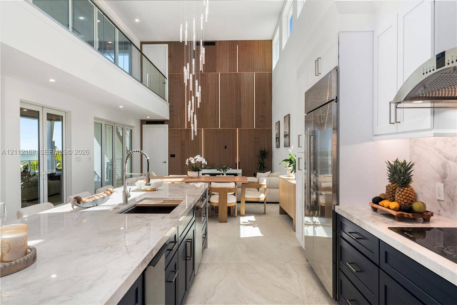 1041 E Buttonwood Drive, Sugarloaf Key, FL 33042 Double-height ceilings, floor-to-ceiling wood-panel accent wall, dual-tone cabinetry with stone-finish countertops, stainless steel integrated appliances, and glass-railing mezzanine