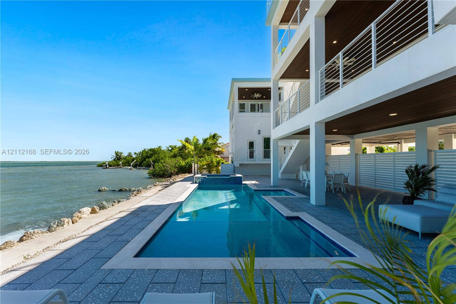 1041 E Buttonwood Drive, Sugarloaf Key, FL 33042 Waterfront exterior featuring a multi-level structure with recessed balconies, horizontal cable railings, a rectangular swimming pool with an integrated spa, and expansive stone-tiled patio decking