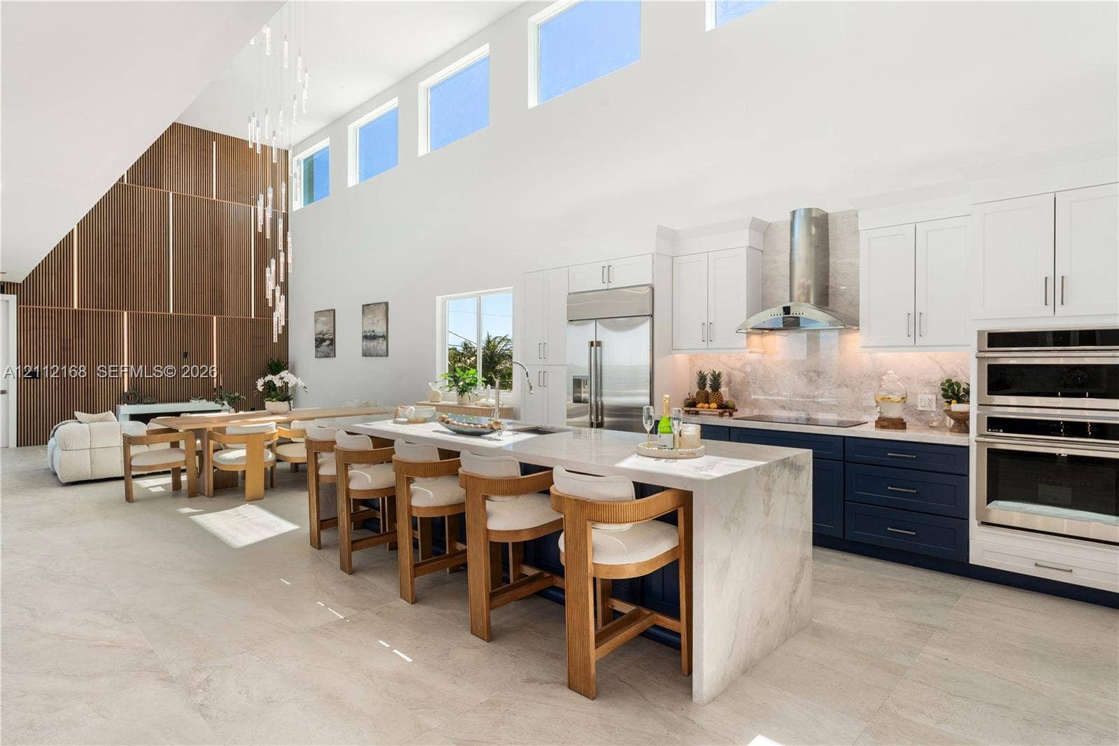 1041 E Buttonwood Drive, Sugarloaf Key, FL 33042 Open-concept kitchen featuring double-height ceilings, a large stone-topped waterfall island, custom two-toned cabinetry, built-in stainless steel appliances, and a floor-to-ceiling wood panel accent wall