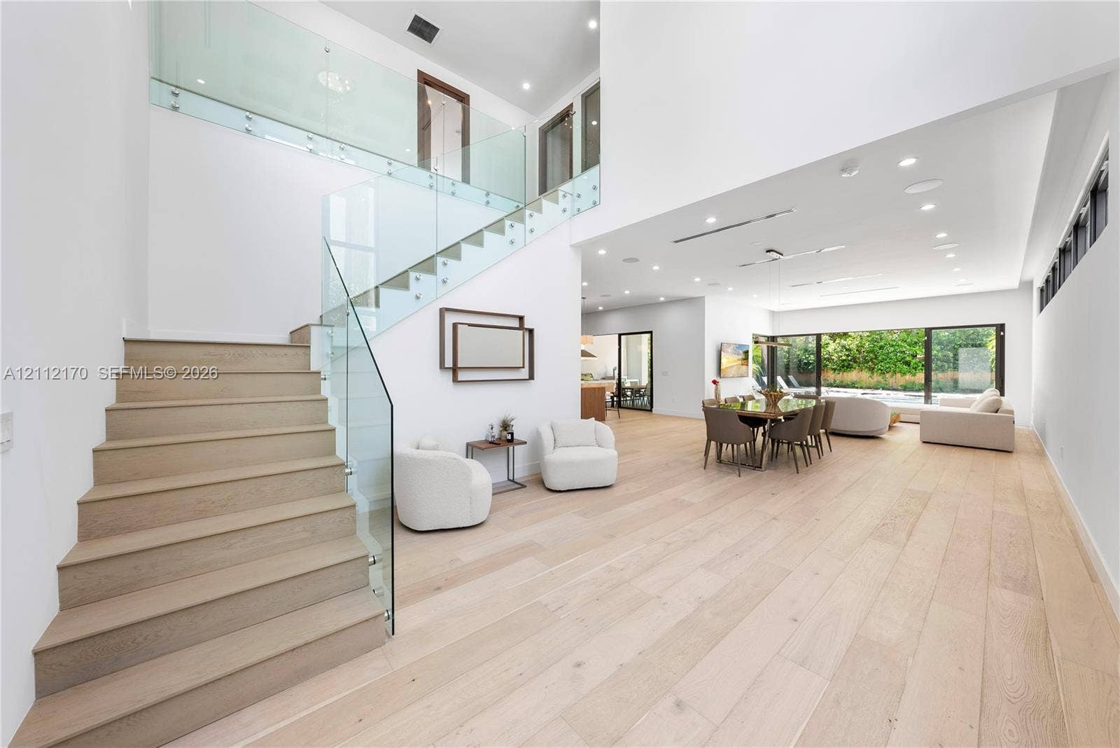 Open-concept interior featuring light wood-finish flooring, staircase with glass railings, recessed lighting, and expansive floor-to-ceiling glass sliding doors