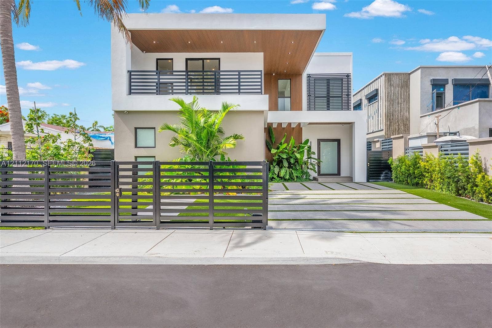 Modern architectural exterior featuring a concrete and wood-paneled facade, second-story balcony with horizontal slat railing, paver driveway with integrated grass strips, and a perimeter metal fence