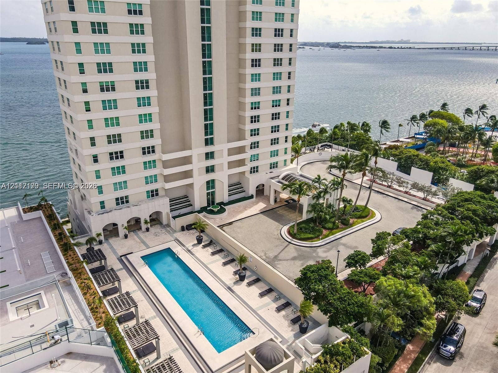 800 Claughton Island Drive, #1604, Miami, Miami, FL 33131 High-rise residential building featuring an expansive outdoor swimming pool, paved pool deck with shade structures, porte-cochre entry, and surrounding mature landscaping, positioned on a waterfront site