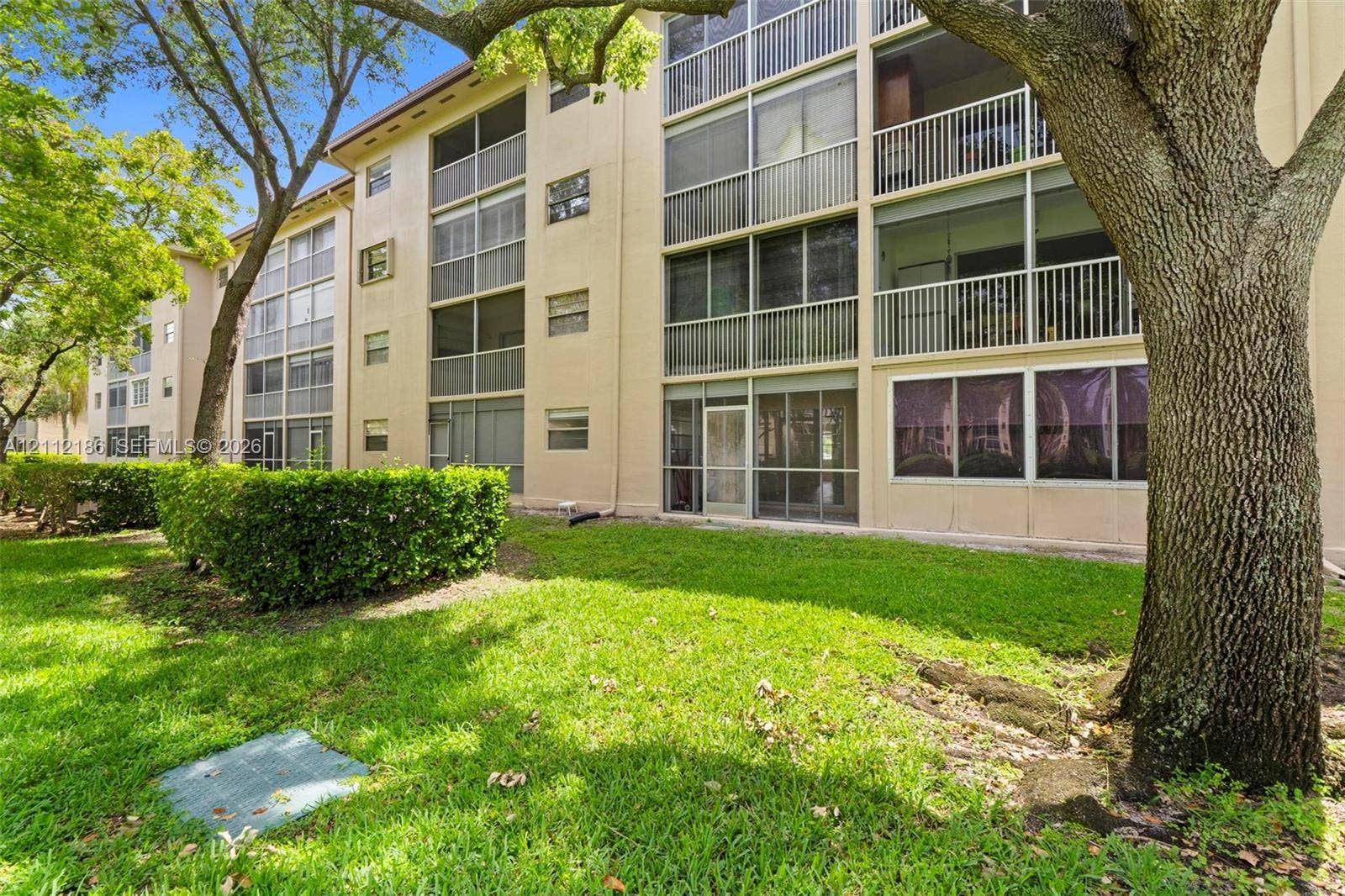1300 SW 124th Terrace, #107P, Pembroke Pines, Pembroke Pines, FL 33027 Multi-story building exterior featuring screened-in lanais, vertical sliding windows, light-toned stucco facade, and manicured landscaping with mature trees