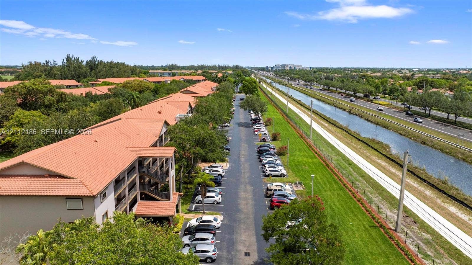 1300 SW 124th Terrace, #107P, Pembroke Pines, Pembroke Pines, FL 33027 Aerial perspective featuring a multi-story building with clay-tile roofing, dedicated asphalt parking, and expansive grassy grounds bordering a waterway