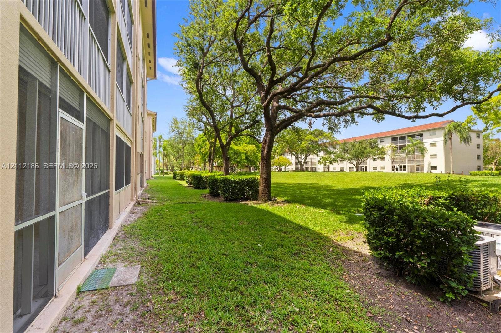 1300 SW 124th Terrace, #107P, Pembroke Pines, Pembroke Pines, FL 33027 Exterior ground-level screen enclosure with full-length window panes, expansive green space, manicured perimeter hedges, and mature canopy trees