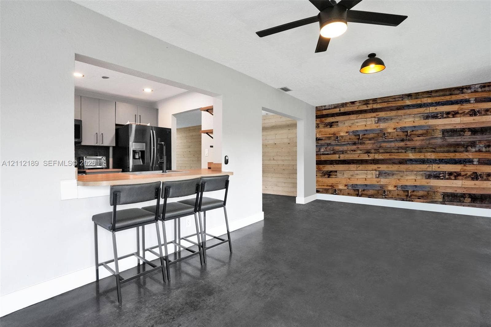 5161 SW 7th Court, #0, Margate, Margate, FL 33068 Interior space featuring a dark polished concrete floor, a horizontal wood-plank accent wall, a kitchen pass-through with a breakfast bar, and a ceiling-mounted matte black fan with integrated lighting
