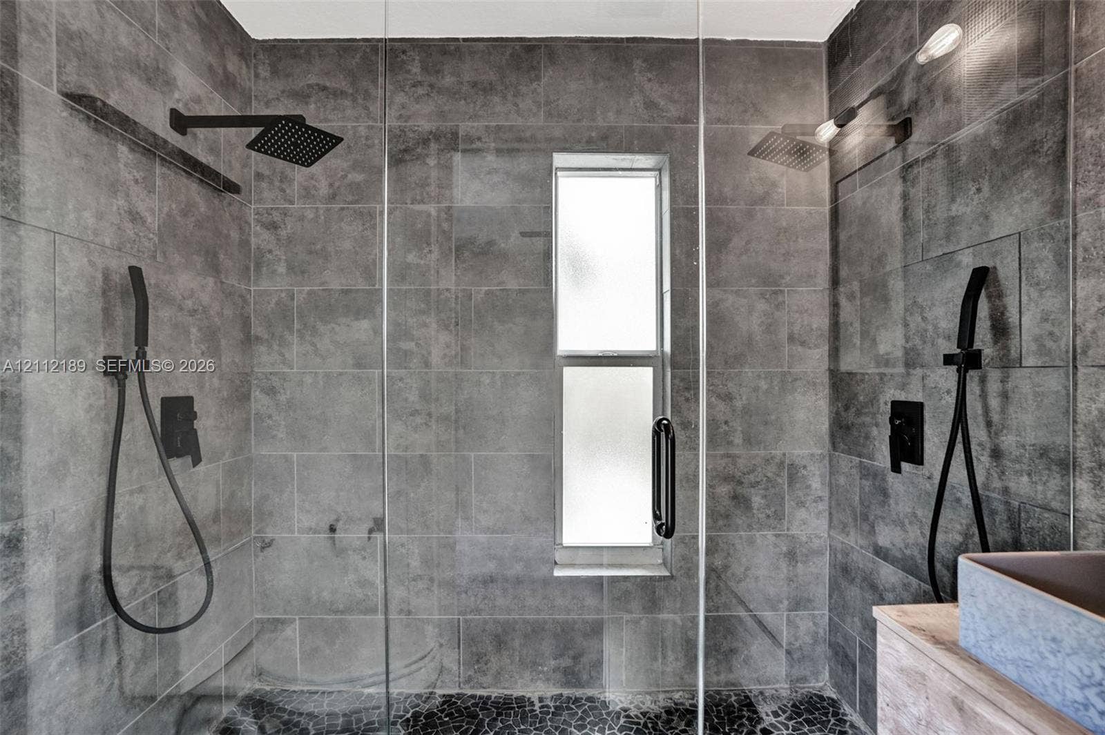 5161 SW 7th Court, #0, Margate, Margate, FL 33068 Walk-in shower featuring large-format grey stone wall tile, black matte rainfall showerheads, handheld shower fixtures, a glass door enclosure, and a stone-tiled floor