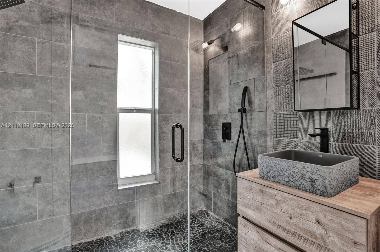 5161 SW 7th Court, #0, Margate, Margate, FL 33068 Bathroom featuring a walk-in shower with floor-to-ceiling textured tile, a frameless glass enclosure, pebble-stone flooring, a wood-finish vanity with a stone vessel sink, and a framed mirror