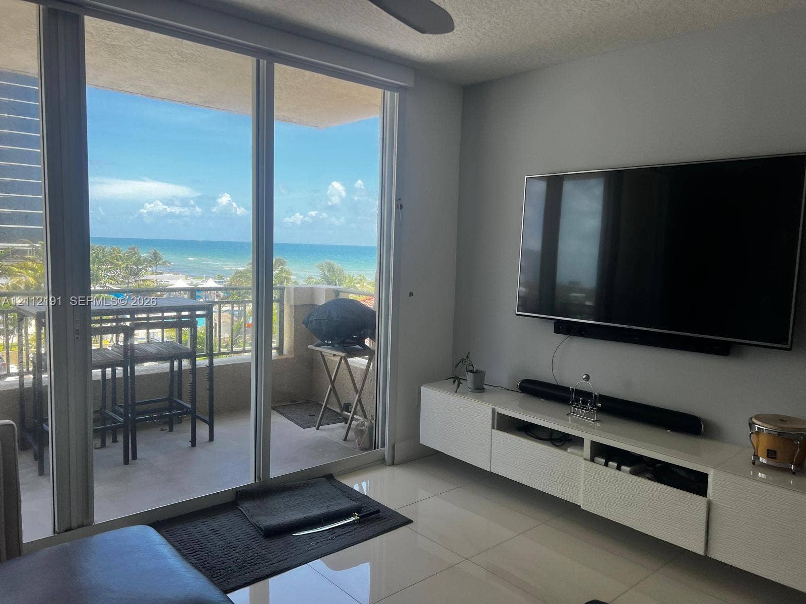 Living area featuring floor-to-ceiling glass sliding doors, stone-finish tile flooring, a wall-mounted floating media cabinet, and a covered balcony with a coastal backdrop