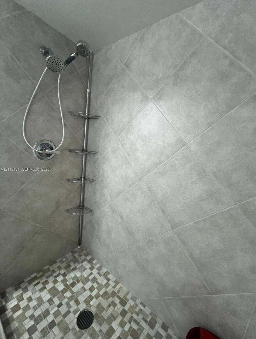 Shower enclosure featuring large-format diagonal wall tiling, mosaic floor tile, multi-setting handheld shower head, and a vertical stainless steel corner shelving unit