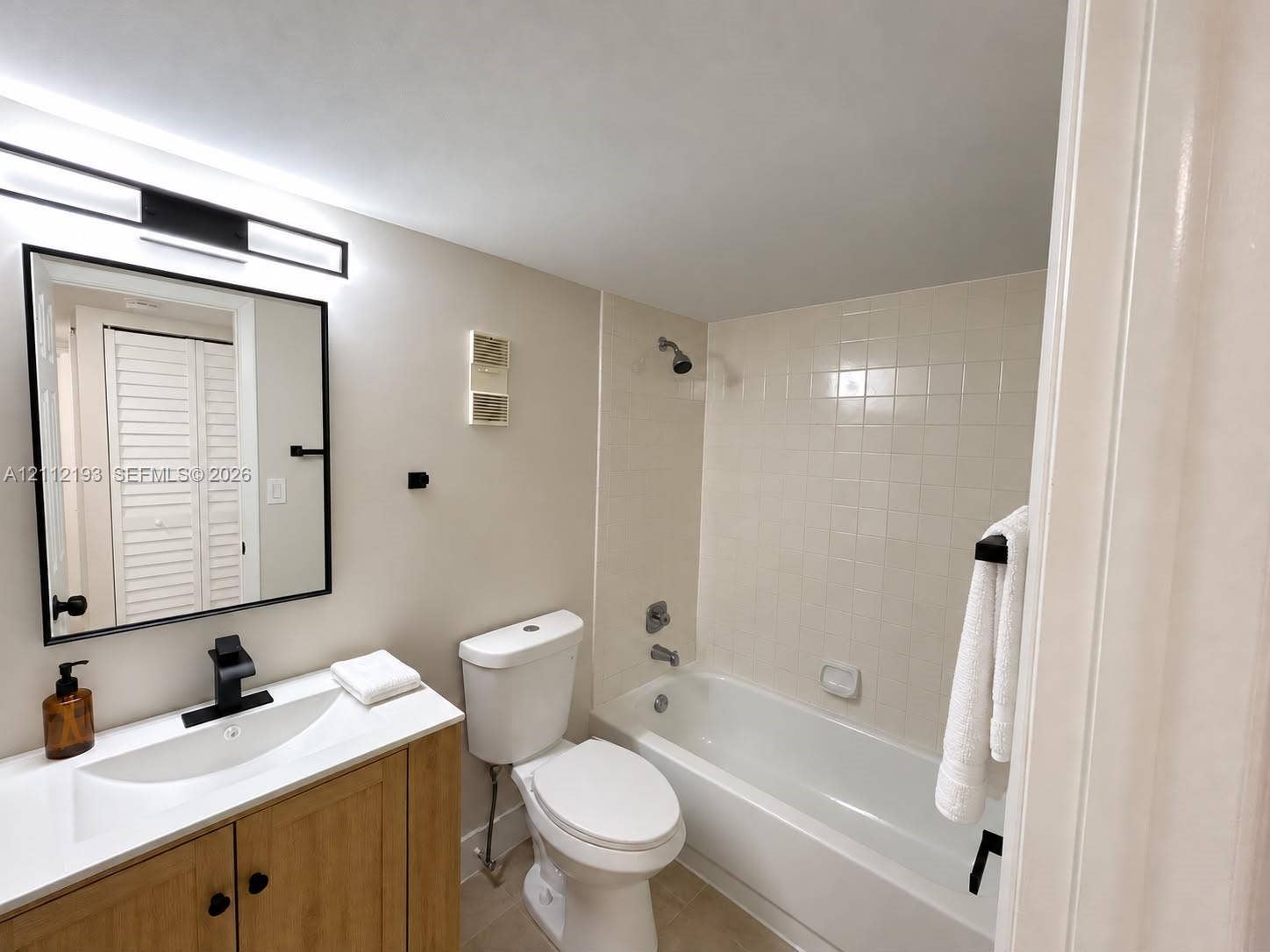 10013 Winding Lake Road, #106, Sunrise, Sunrise, FL 33351 Bathroom featuring a wood-finish vanity with a white integrated sink, matte black faucet, rectangular framed mirror with integrated lighting, and a tiled shower-tub combination