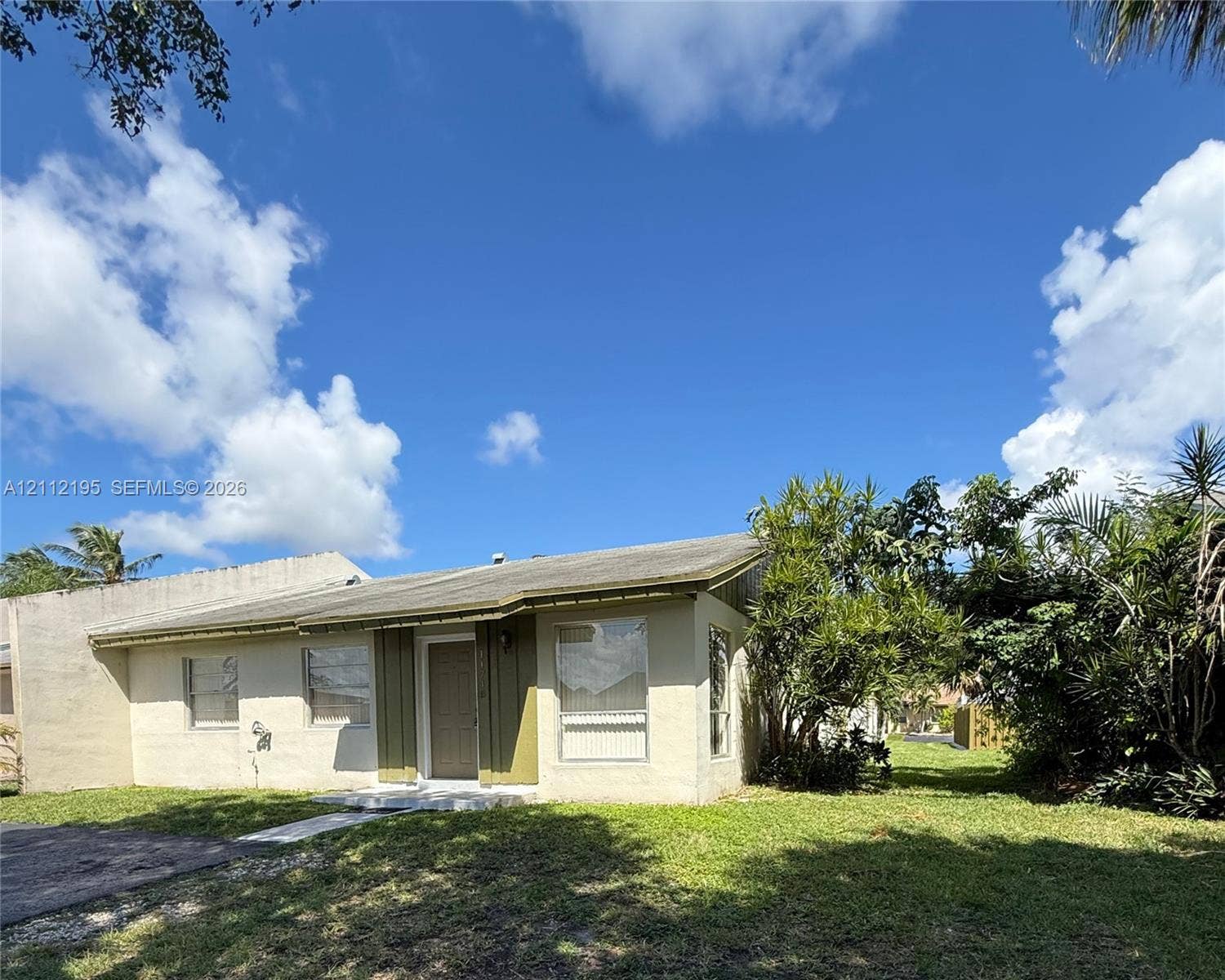 11745 SW 122nd Place, #11745, Miami, Miami, FL 33186 Single-story residence featuring a low-slope roof, light-toned stucco exterior, vertical paneling at the entryway, and expansive grassy grounds with mature trees