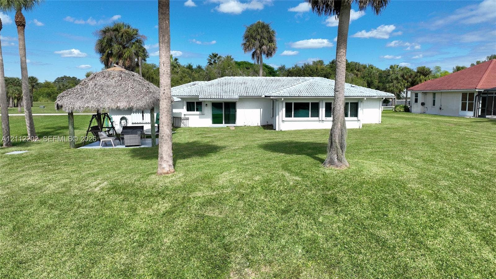 3233 Shell Lane, Labelle, FL 33935 Rear exterior featuring a sprawling lawn, a detached thatched-roof palapa, and mature palm trees