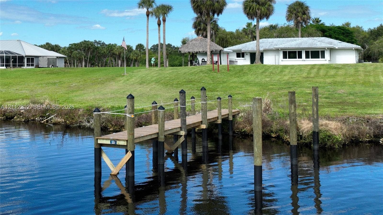 3233 Shell Lane, Labelle, FL 33935 Waterfront property featuring a private wooden dock, sprawling grass grounds, mature palm trees, and a detached thatched-roof structure