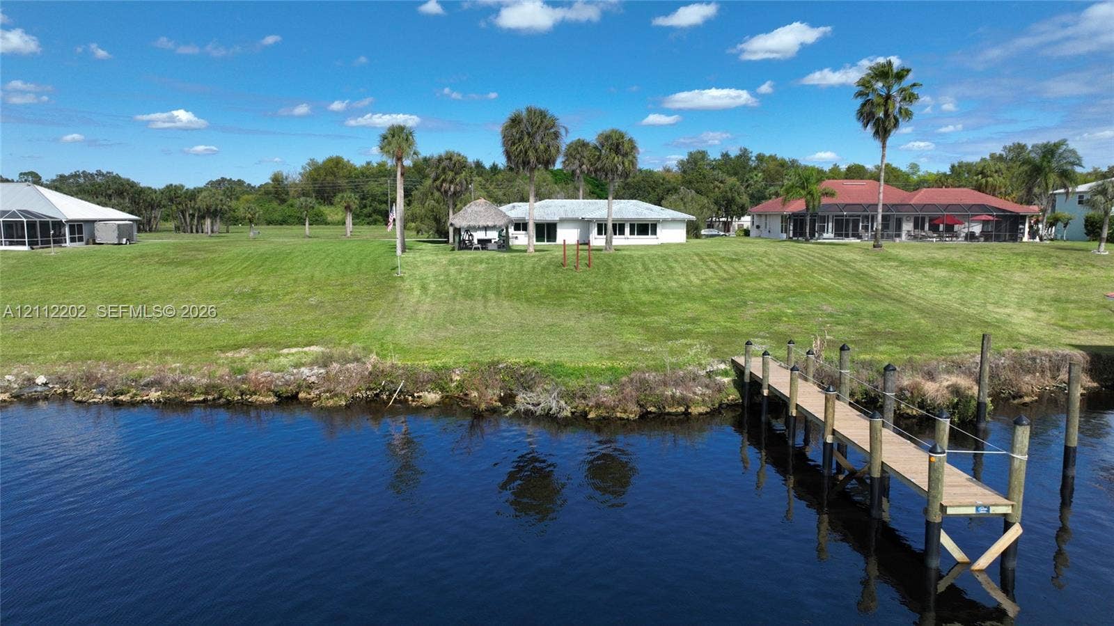 3233 Shell Lane, Labelle, FL 33935 Waterfront property featuring a private wooden dock, expansive lawn, detached thatched-roof structure, and mature palm trees