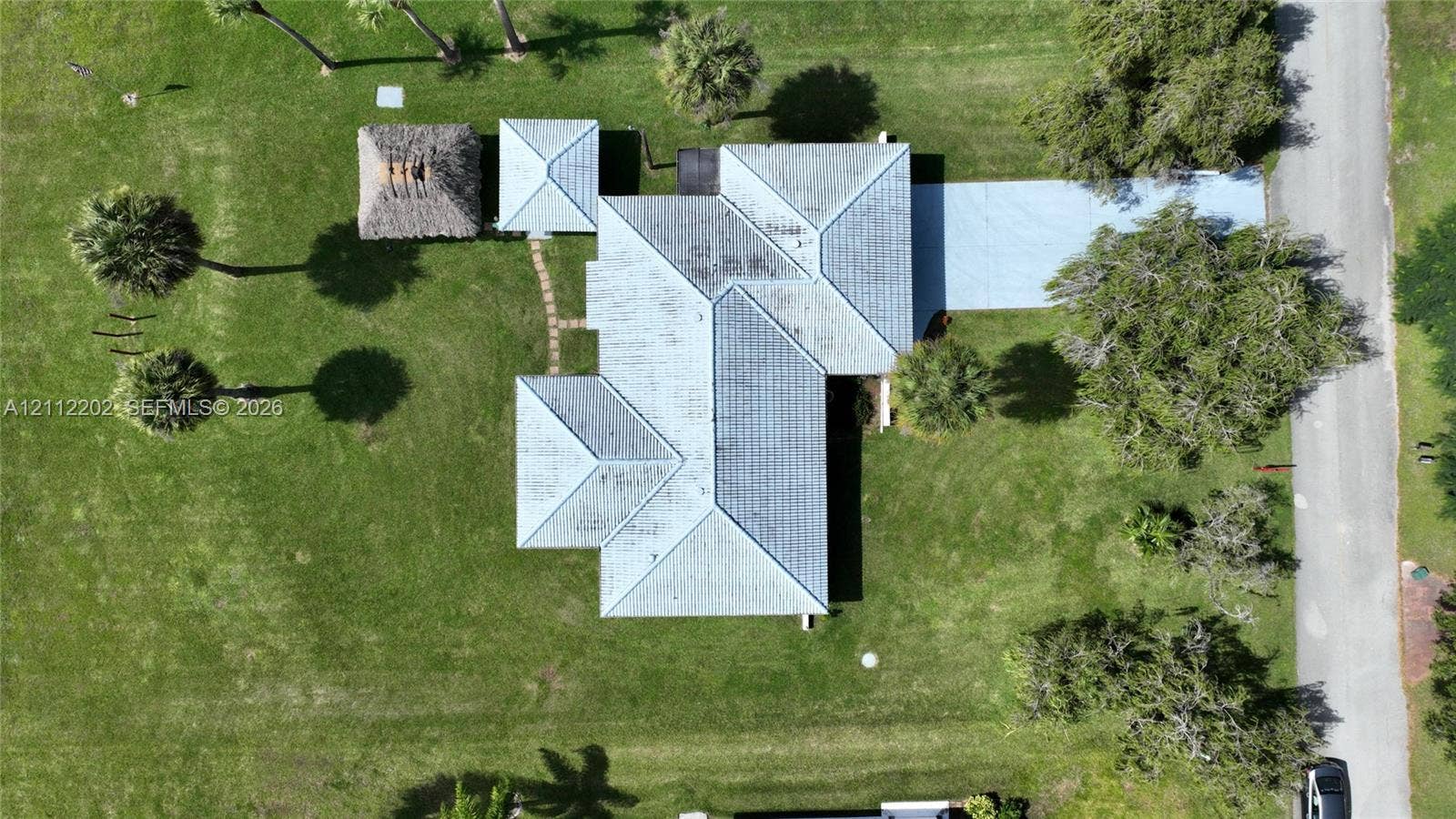 3233 Shell Lane, Labelle, FL 33935 Aerial perspective of property featuring a multi-pitched metal roof, paved driveway, detached thatched-roof structure, and expansive grass lawn with mature palm trees