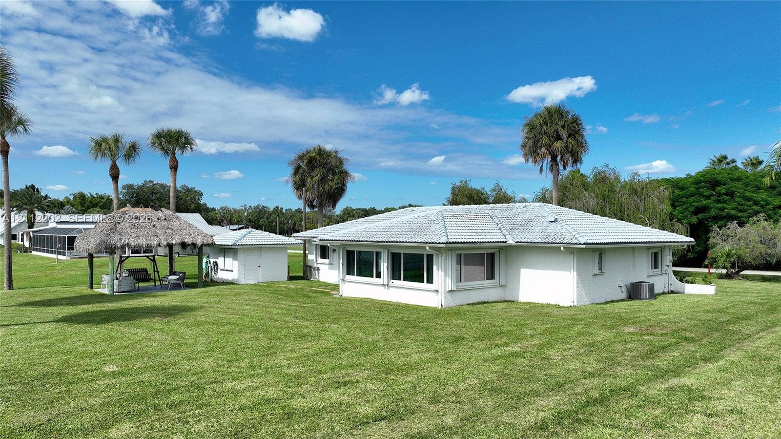 3233 Shell Lane, Labelle, FL 33935 Exterior featuring white painted masonry, a hipped roof with light-colored tile, a thatched-roof structure, and an expansive lawn with mature palm trees