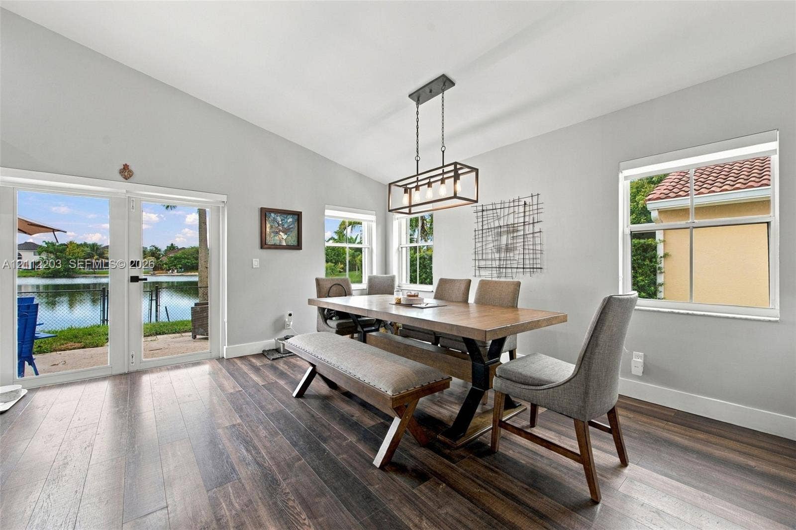 Vaulted ceiling room with wood-finish flooring, linear metal pendant lighting, and glass-paneled double doors providing water frontage