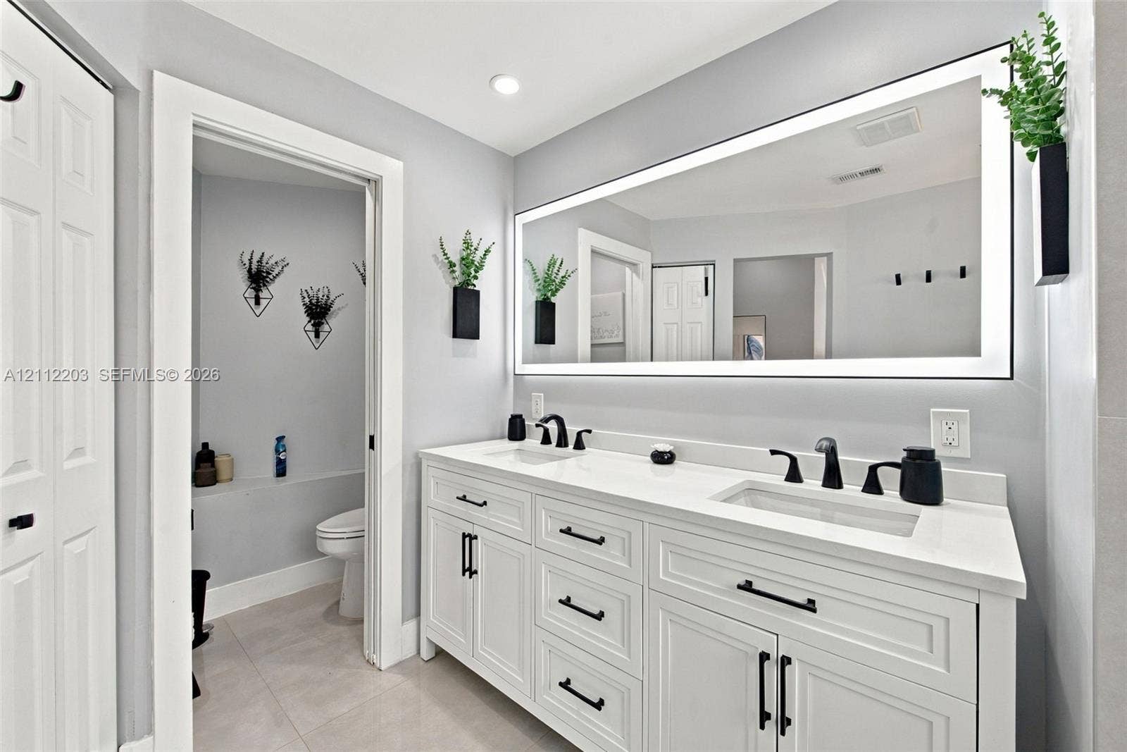 Dual-sink vanity with white cabinetry, light-toned countertop, matte black hardware, and an integrated LED-lit vanity mirror