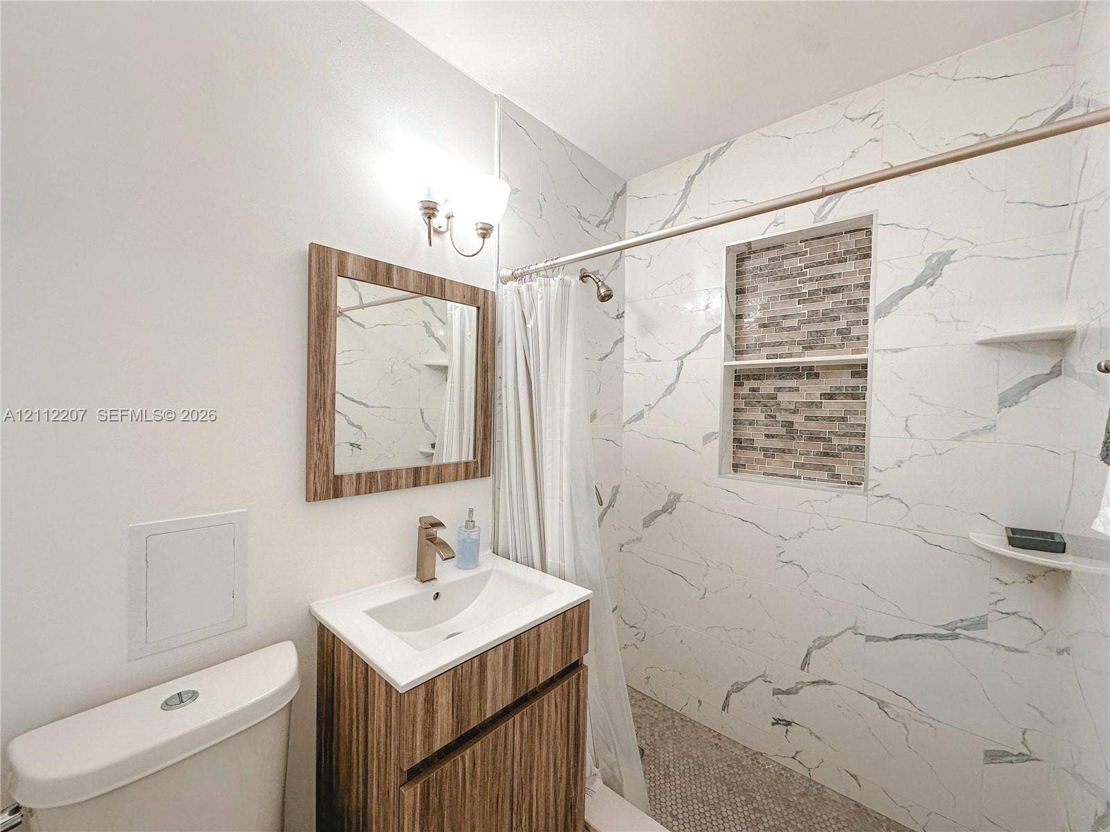 525 SW 67th Avenue, Miami, Miami, FL 33144 Bathroom featuring a vanity with a white countertop, wall-mounted light fixture, mirror with a wood-finish frame, and a shower area with large-format tile surround, stone-finish tile accent niche, and built-in shelving