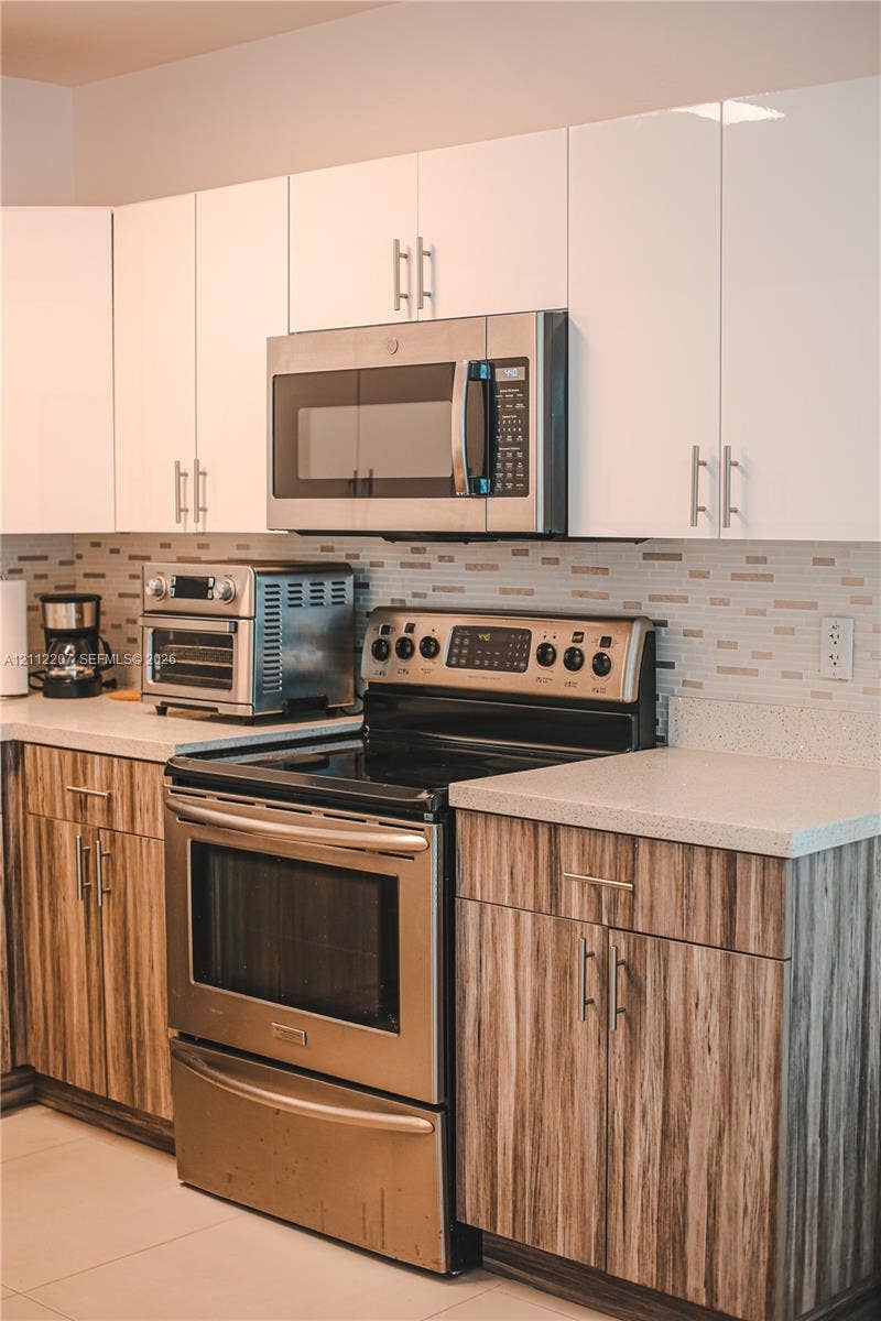 525 SW 67th Avenue, Miami, Miami, FL 33144 Kitchen featuring high-gloss white upper cabinetry, wood-finish lower cabinetry, stone-composite countertops, stainless steel appliances, and a linear tile backsplash